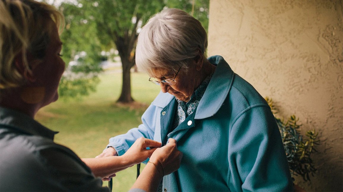 A healthcare worker helps an older adult button her jacket.